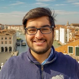 An Indian male smiling at the camera. He has medium-length dark hair, dark-rimmed glasses, and stubble. He's wearing a blue zip-up sweater. In the background is a canal lined with old-looking white and orange buildings.